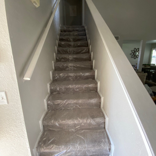 Staircase with carpet covered in plastic for protection, leading upwards. White banister and gray walls.