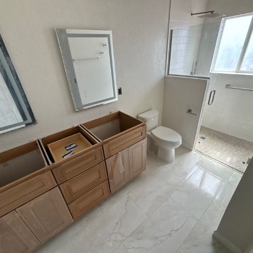 Bathroom renovation in progress; cabinets installed, toilet and shower visible, white tile floor, large mirror.
