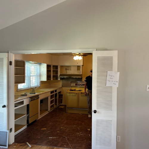 Kitchen with cabinets, oven, and dishwasher. A person stands in the kitchen doorway. Interior room with brown floors.