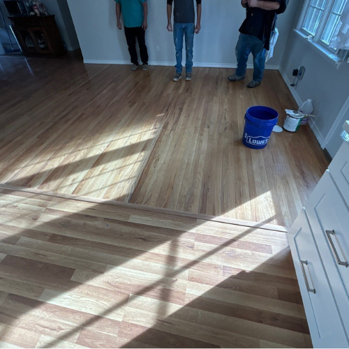 Three people standing on a newly installed hardwood floor. Blue bucket and supplies are nearby.