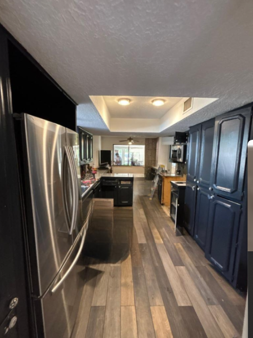 Narrow kitchen with stainless fridge, dark cabinets, island, and wood-look floor under recessed lights