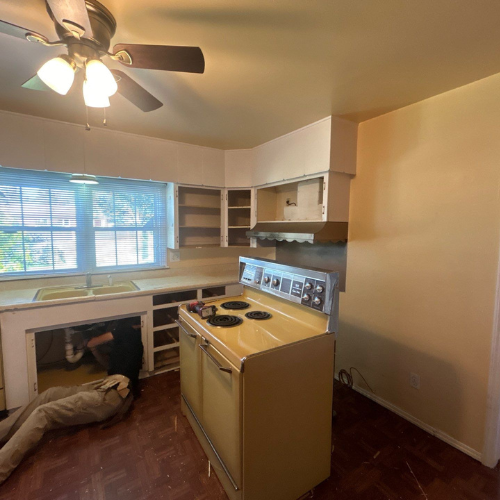Kitchen renovation: person working under sink; yellow vintage stove, empty cabinets, brown ceiling fan.