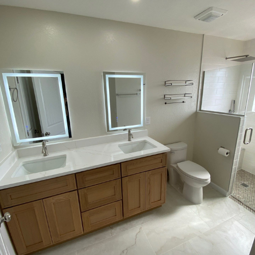 Bathroom with double vanity, illuminated mirrors, toilet, and glass shower.