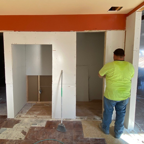 Person in neon shirt working on drywall in a room under construction; orange wall, unfinished floors.