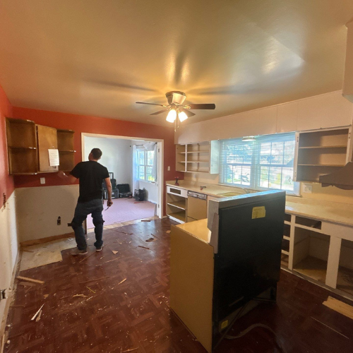 Man works on kitchen remodel with cabinets removed. Red walls, brown floor, open to living room.