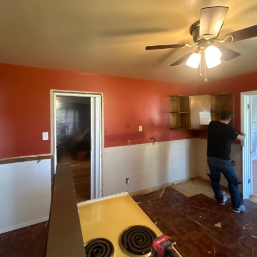Kitchen renovation: Person removing cabinet from red wall; tan walls below; brown floor; off-white doors; yellow stove.