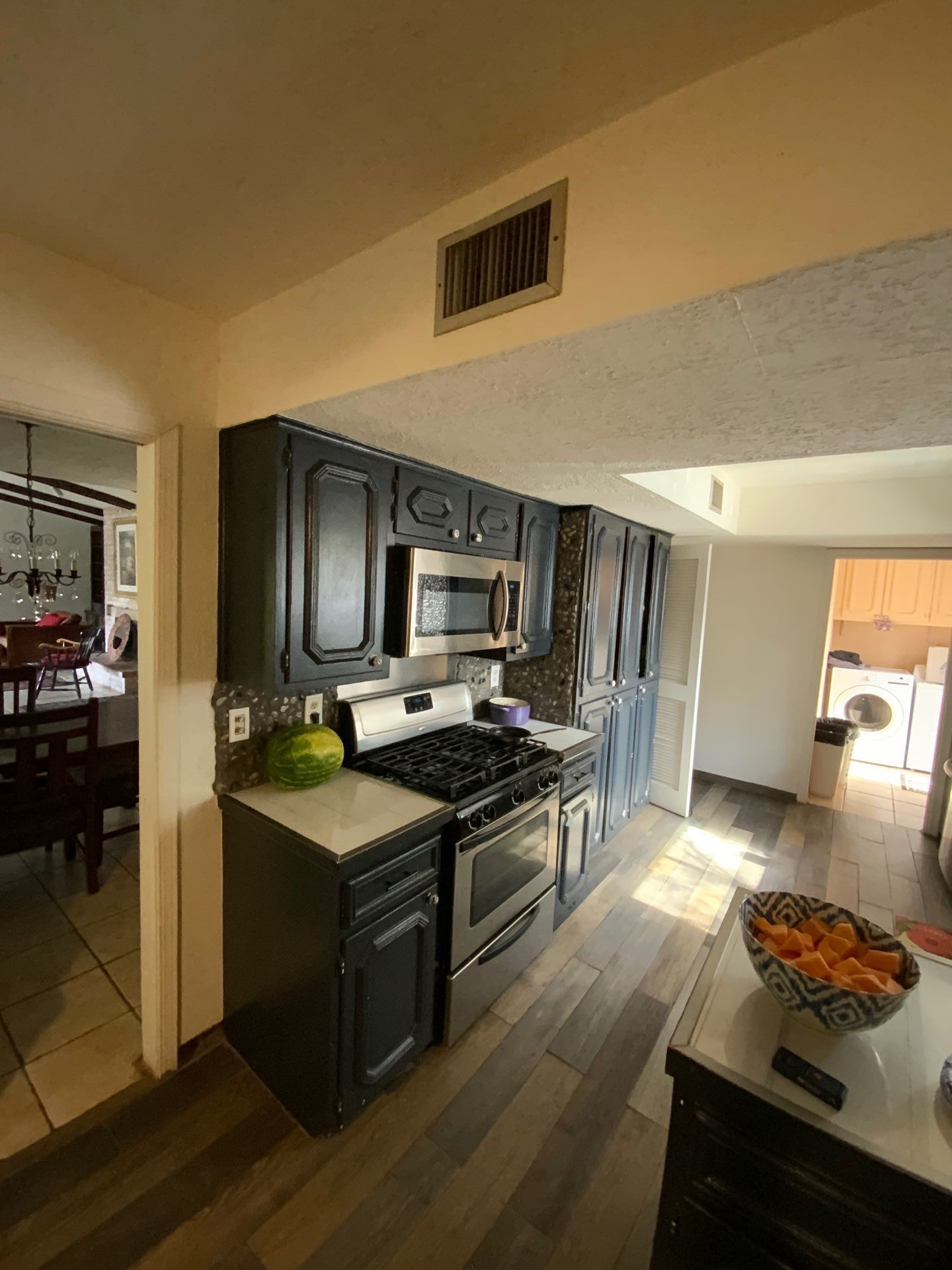 Kitchen with dark cabinets, stainless stove, and a bowl of fruit on an island, viewed into a bright dining area
