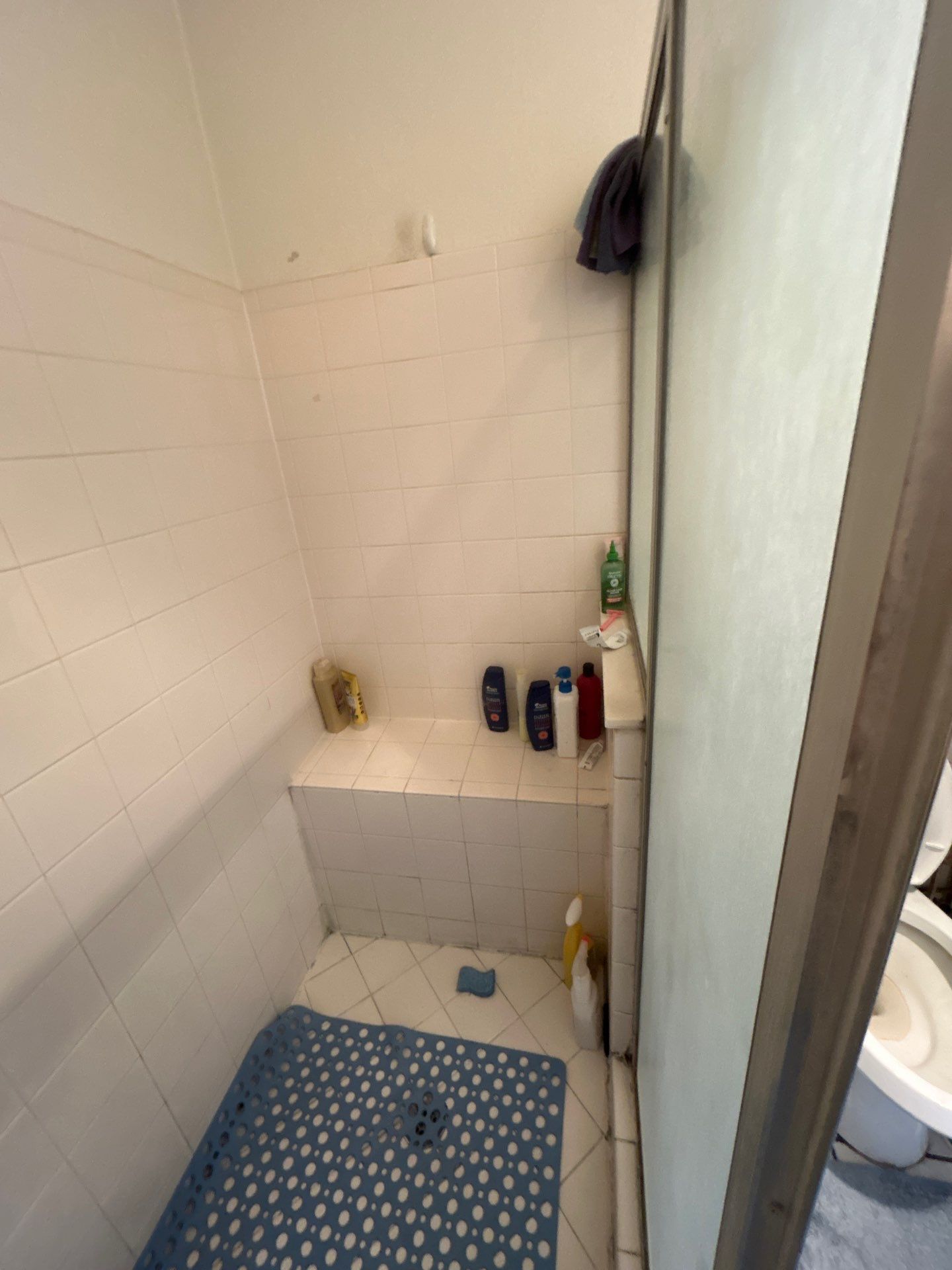 Small white tiled shower with a built-in bench, blue bath mat, and bottles of shampoo and soap.
