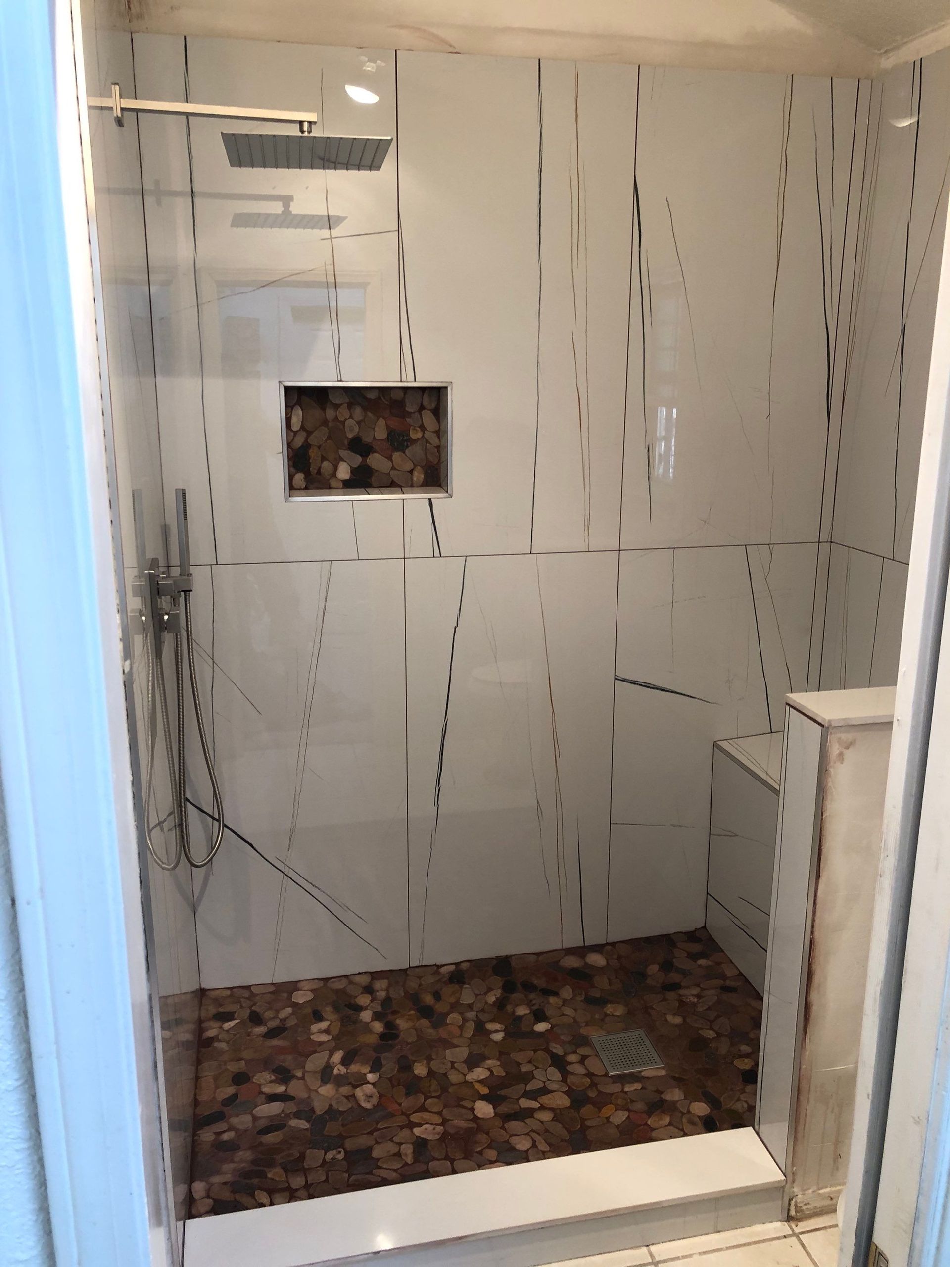 Shower with white walls and a stone floor, featuring a built-in bench and a recessed shelf.