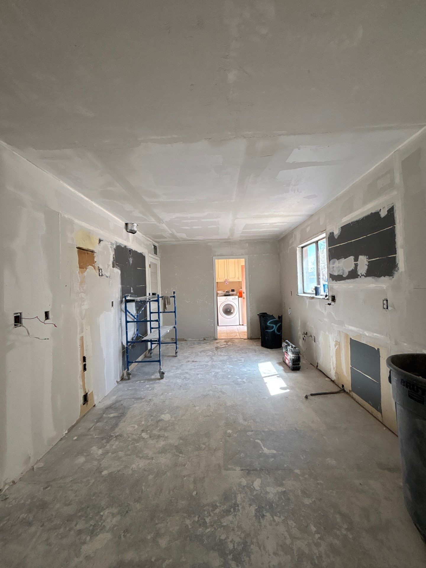 Narrow unfinished hallway with drywall patches, tools, and a window at the far end.