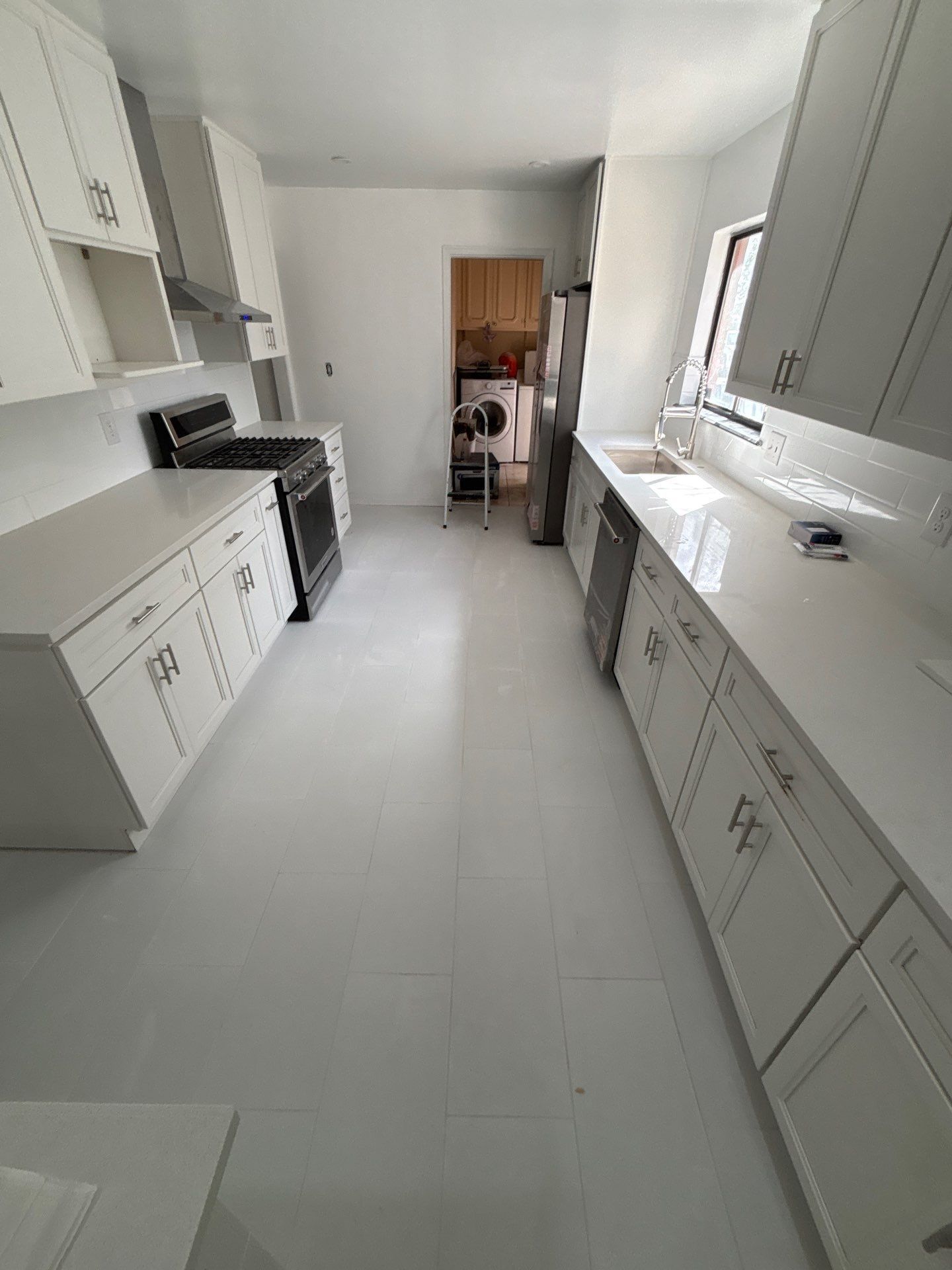Bright narrow white kitchen with long counters, cabinets, and a stove; a person stands at the far end.