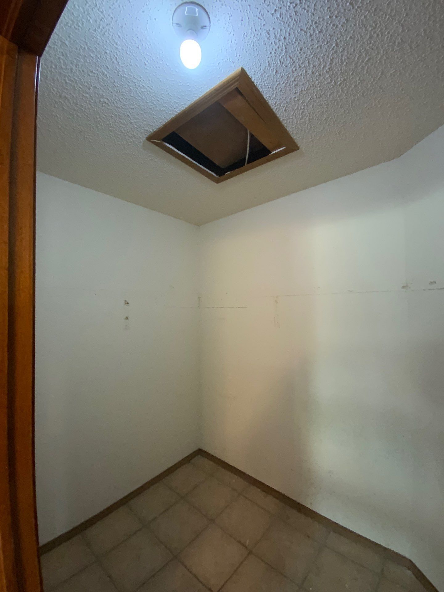 Small room with a square ceiling hatch and a light. Tile floor, white walls. Brown wooden door frame visible.