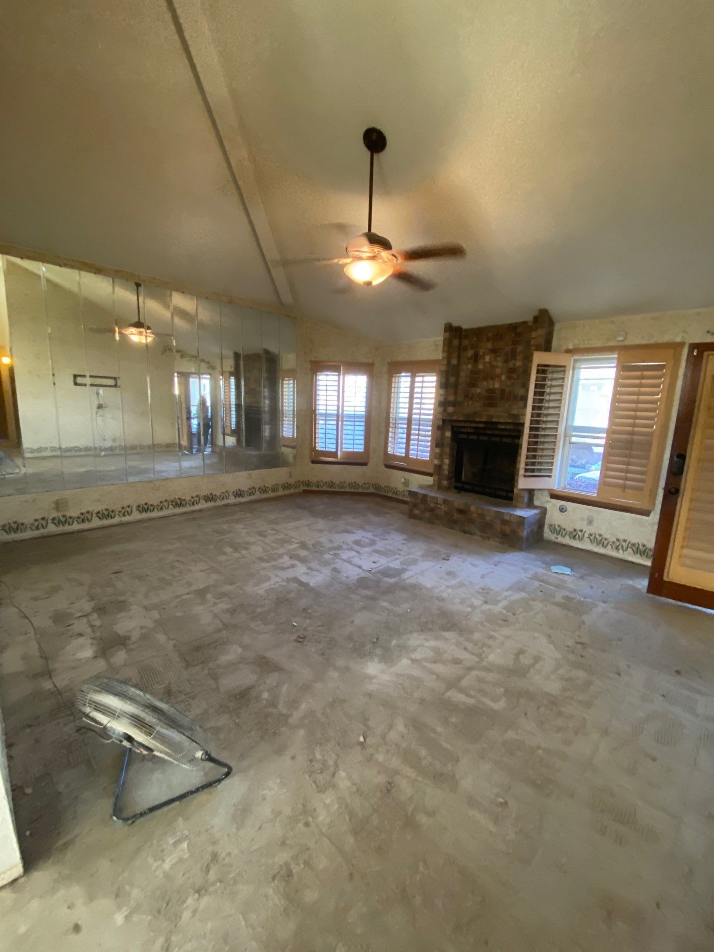 Empty living room with bare concrete floor, fireplace, windows, mirror wall, ceiling fan.