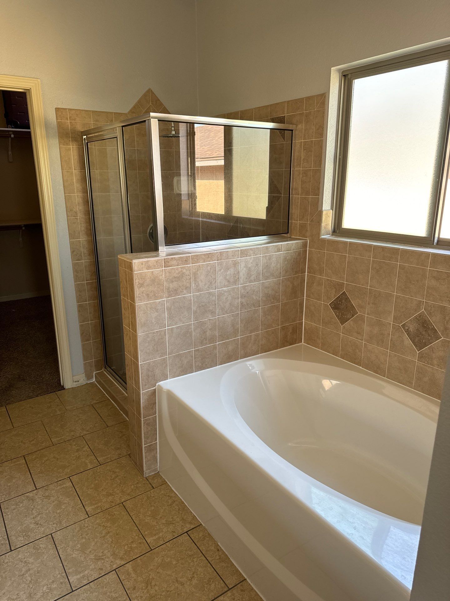 Bathroom with tan tiled walls, white bathtub, glass shower, and window.