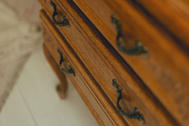 Primo piano angolato di una cassettiera in legno con quattro cassetti e maniglie in metallo decorate, appoggiata su un pavimento bianco.