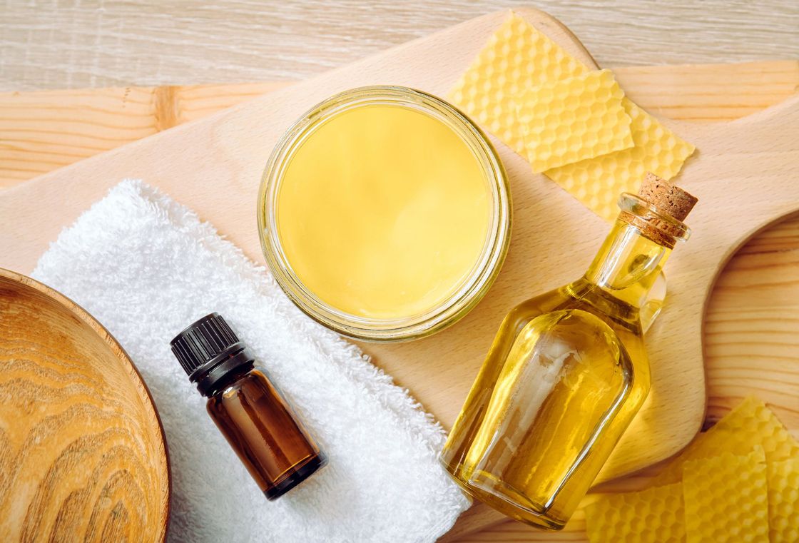 Una superficie di legno cosparsa di cera d'api, un vasetto di balsamo, una bottiglia di vetro di olio, un contagocce per oli essenziali e un asciugamano bianco.