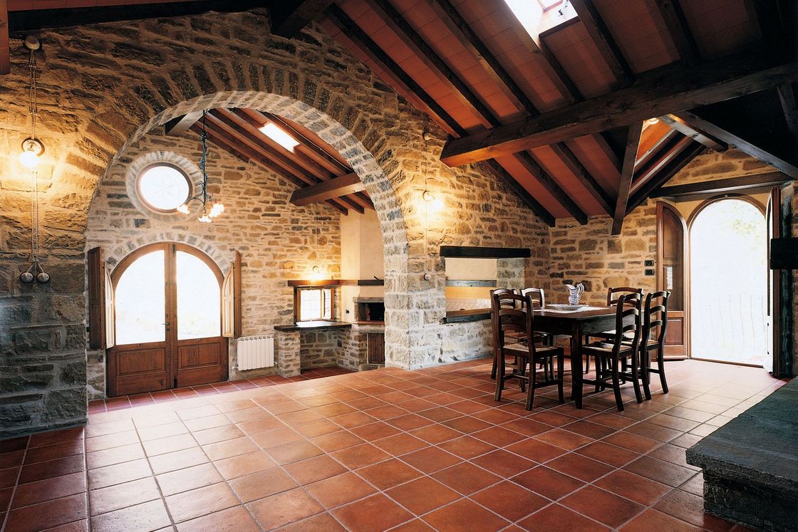Una stanza rustica caratterizzata da pareti in pietra, un ingresso ad arco, travi in ​​legno a vista, pavimento in piastrelle di terracotta e un set da pranzo.