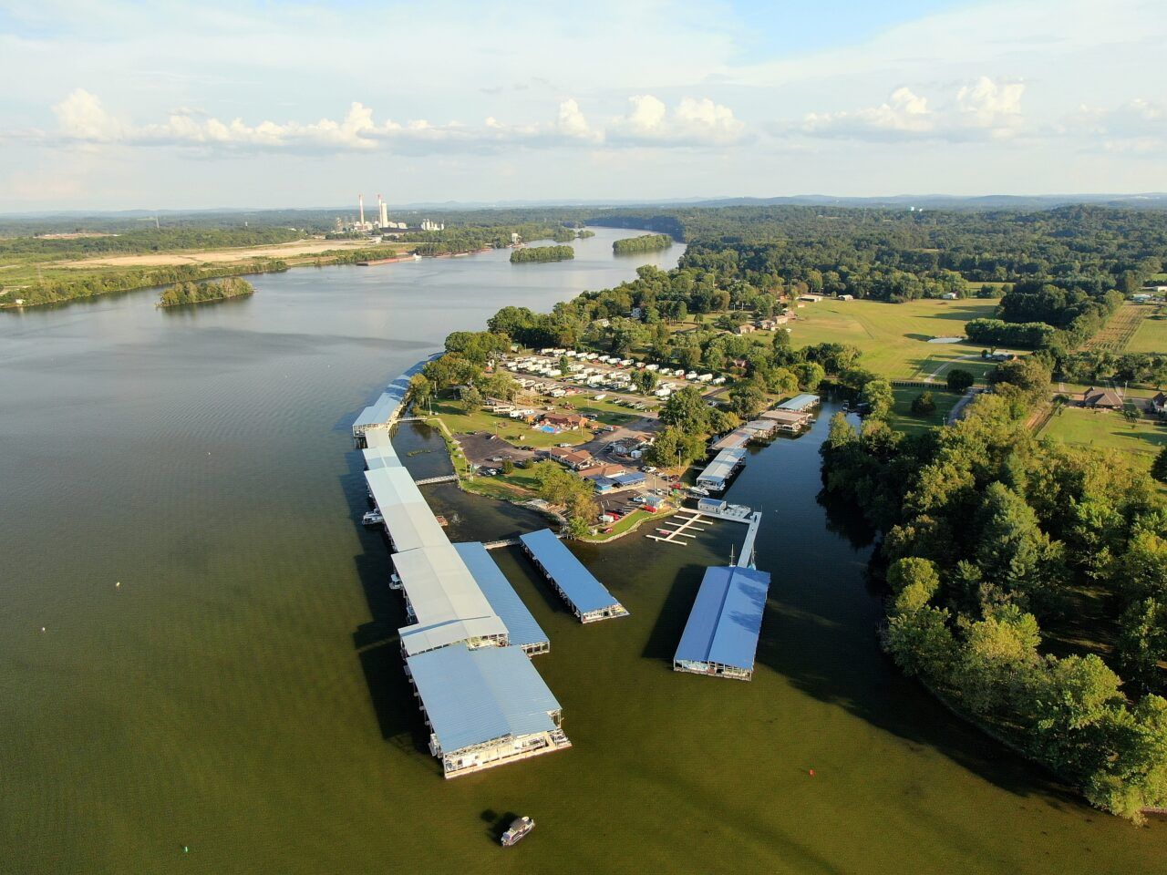 Aerial view of covered boat docks and waterfront cottages on a wide lake, with a power plant in the distance