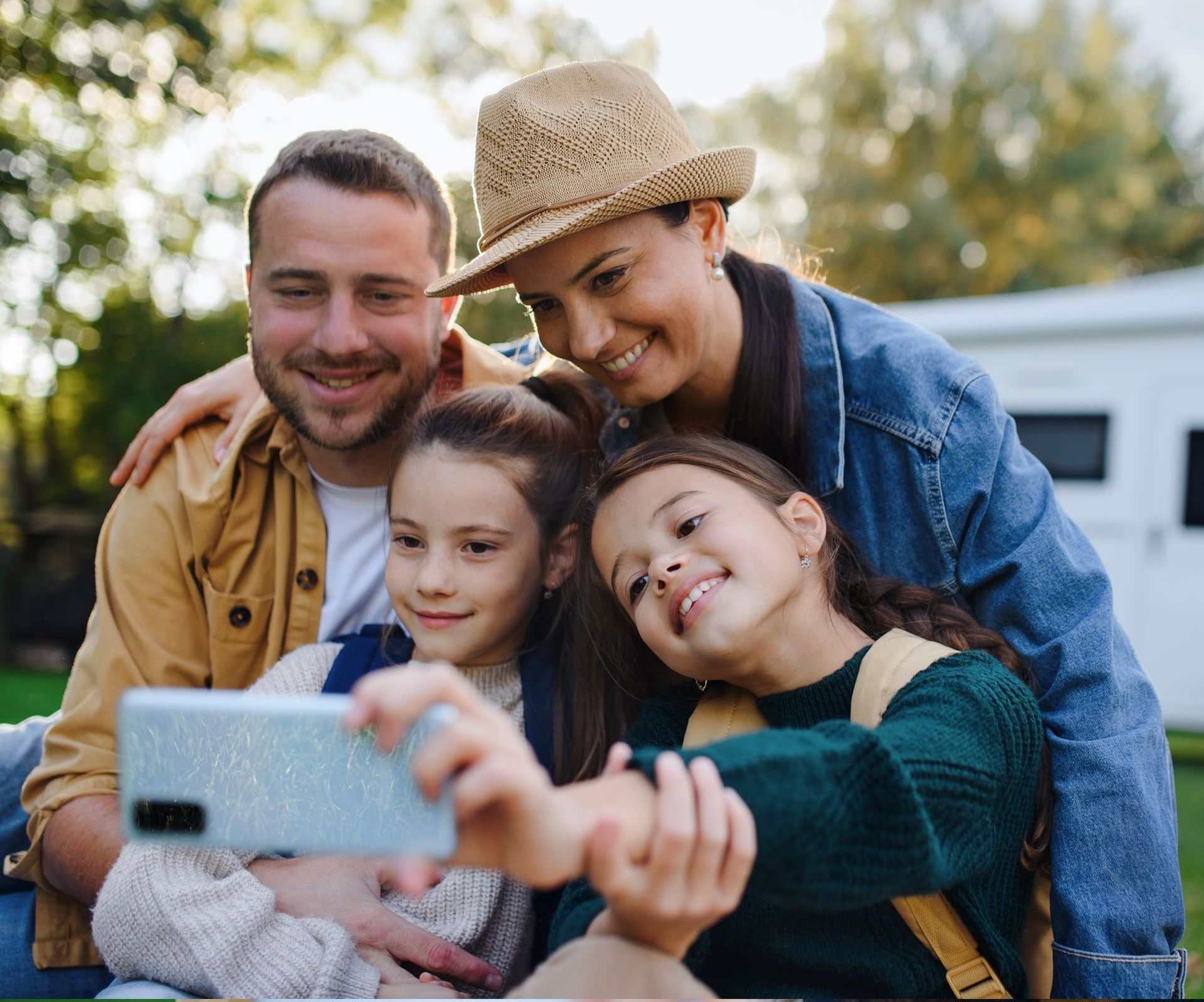 Smiling family of four taking a selfie outdoors, with an RV behind them