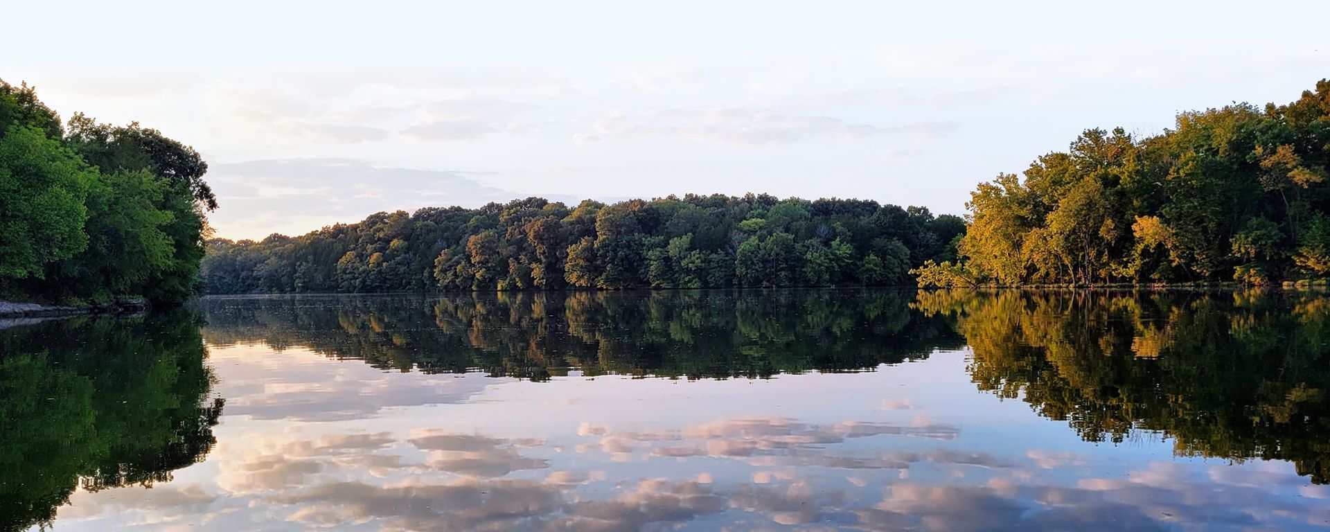 Calm lake reflecting a tree-lined shore and soft clouds at dusk