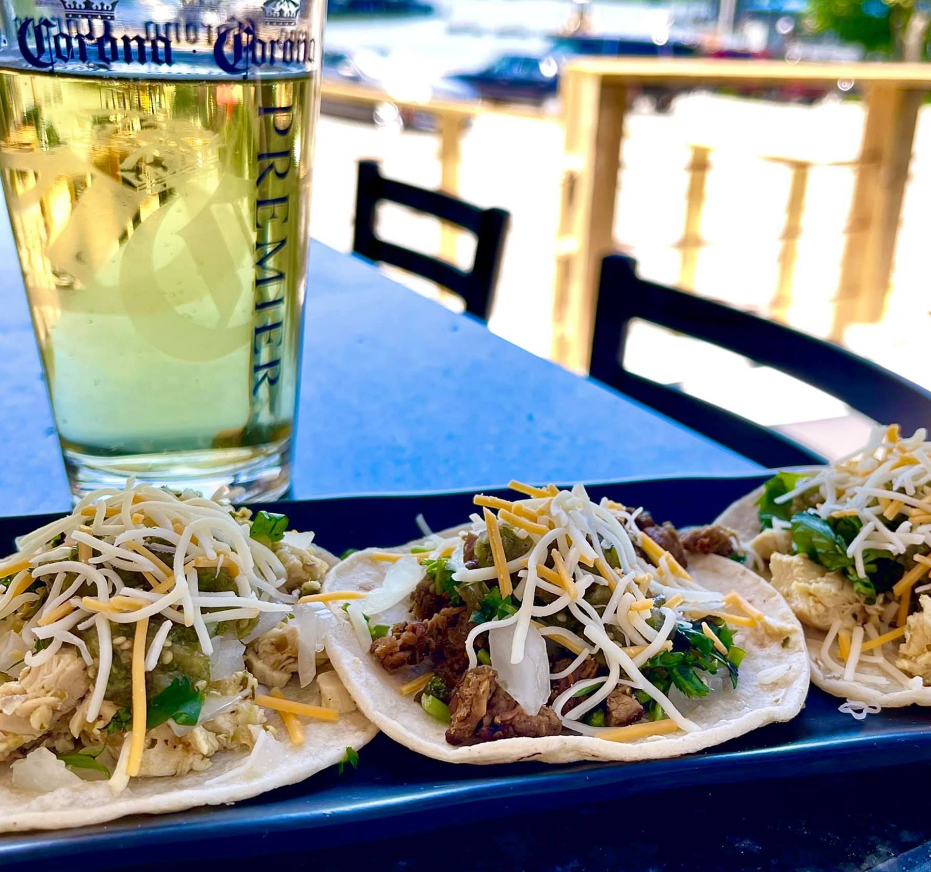Tacos topped with shredded cheese and a glass of beer on a patio table