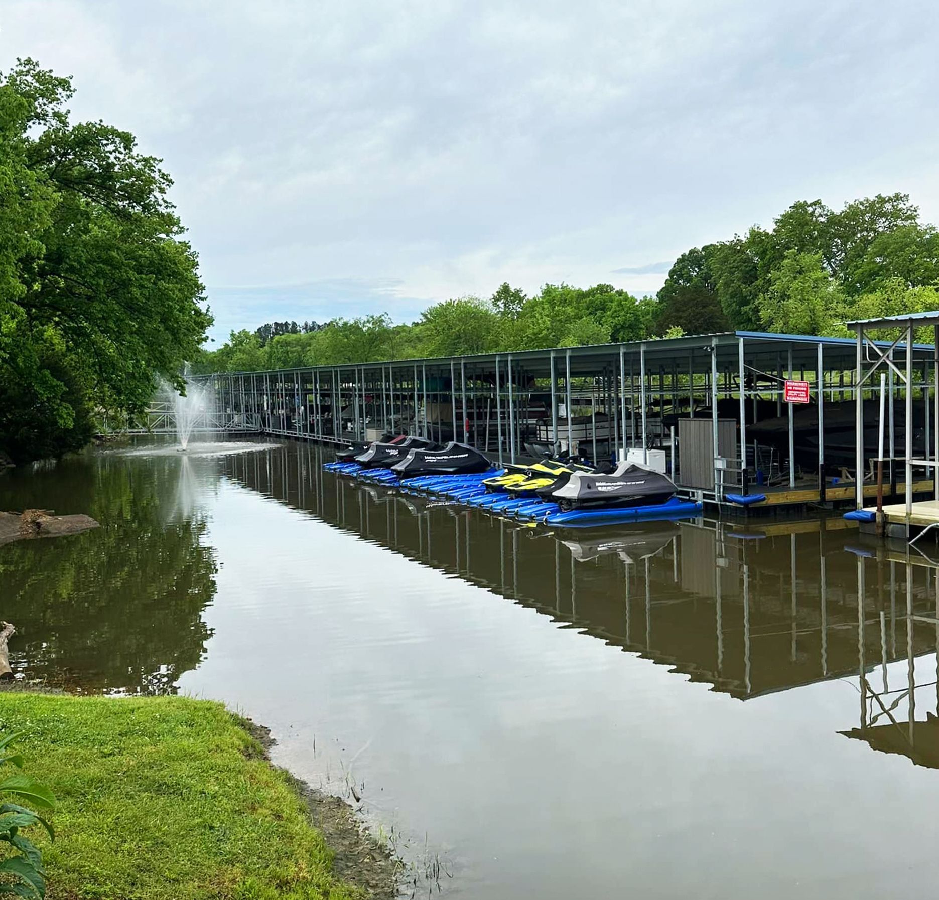 Jet skis parked on blue floating docks beside a covered boat dock, with a fountain on the water