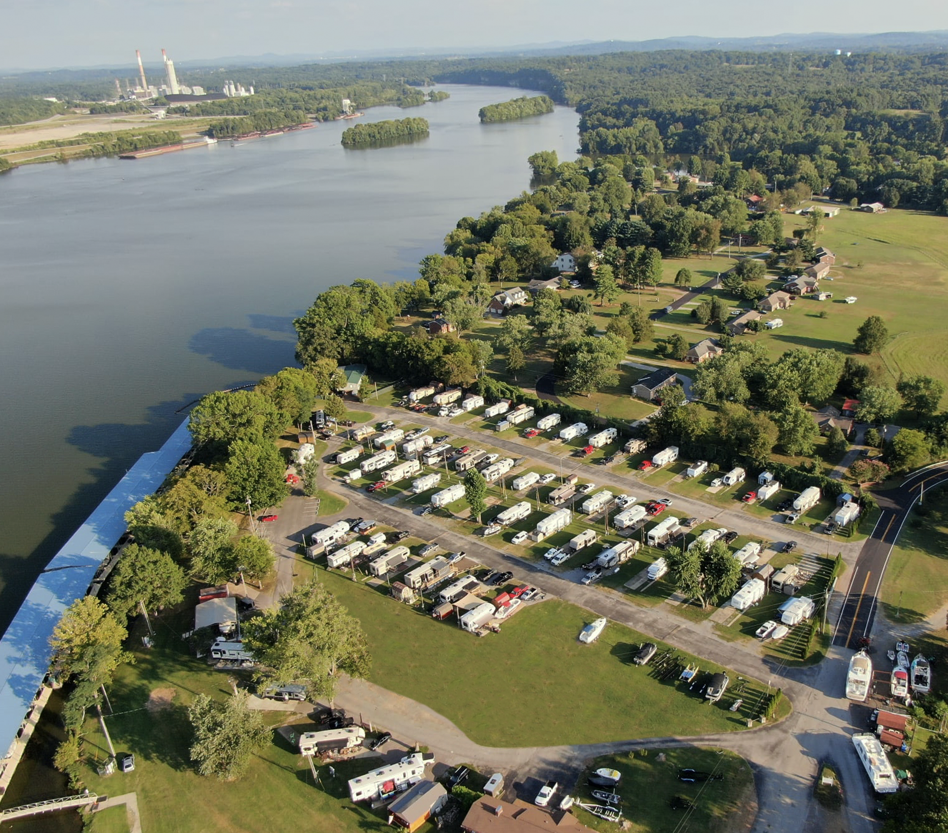Aerial view of an RV park with rows of campers beside a wide river, with a power plant in the distance