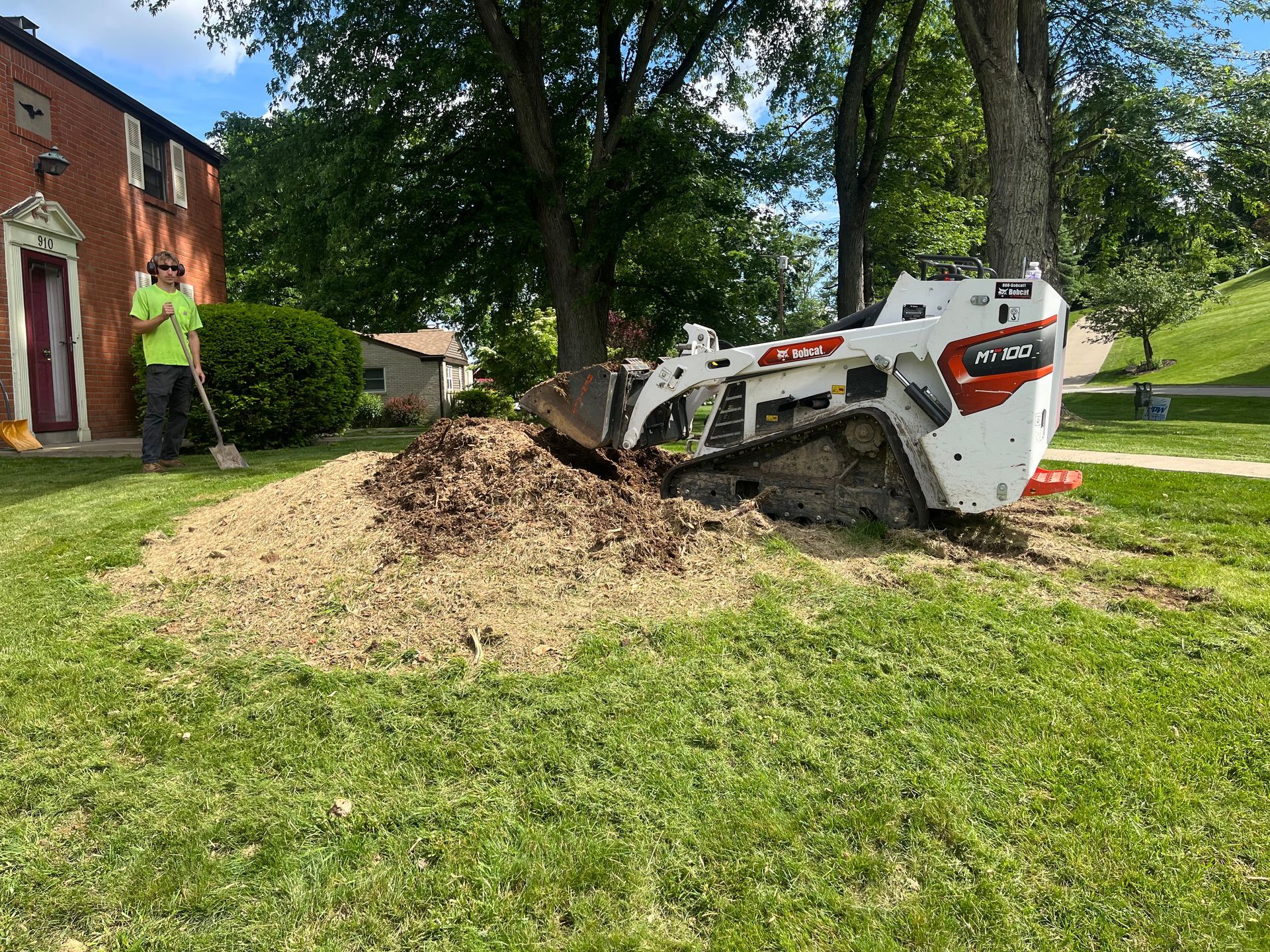 Worker operating a skid steer, digging soil beside a brick house on a sunny lawn.