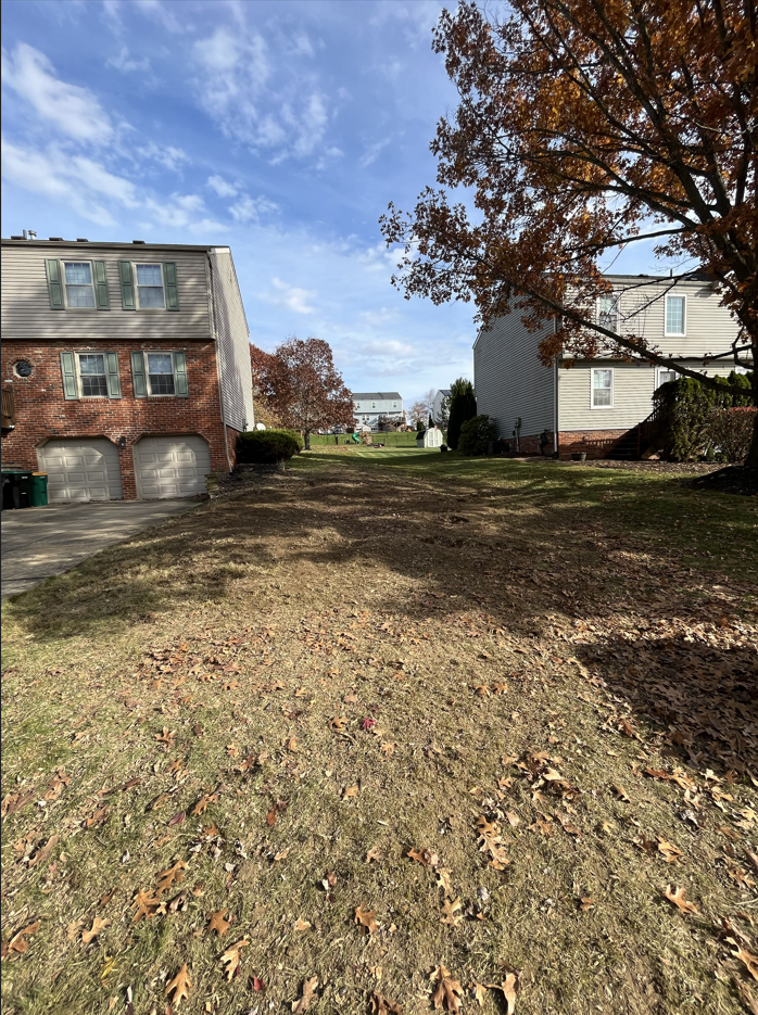 Leaf-covered grassy yard between two houses under a blue sky, with autumn trees and a driveway on the left