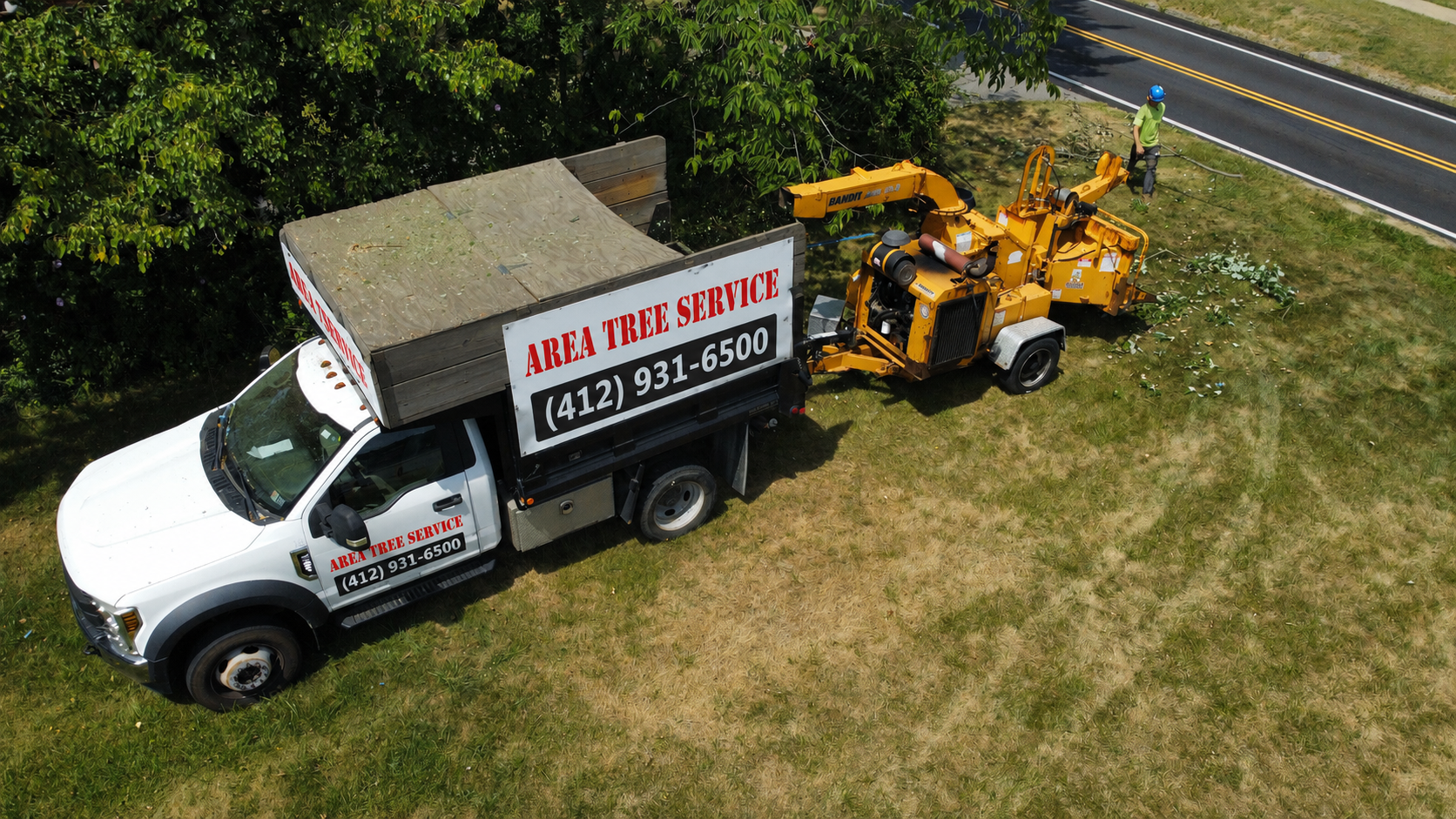 White truck with a sign and yellow digging equipment parked on grass near trees