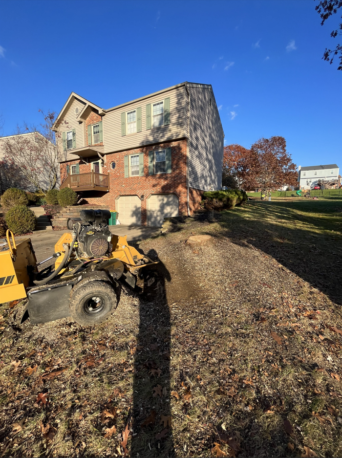 Two-story house with brick and siding, yard equipment in foreground, and a long shadow on a sunny day.