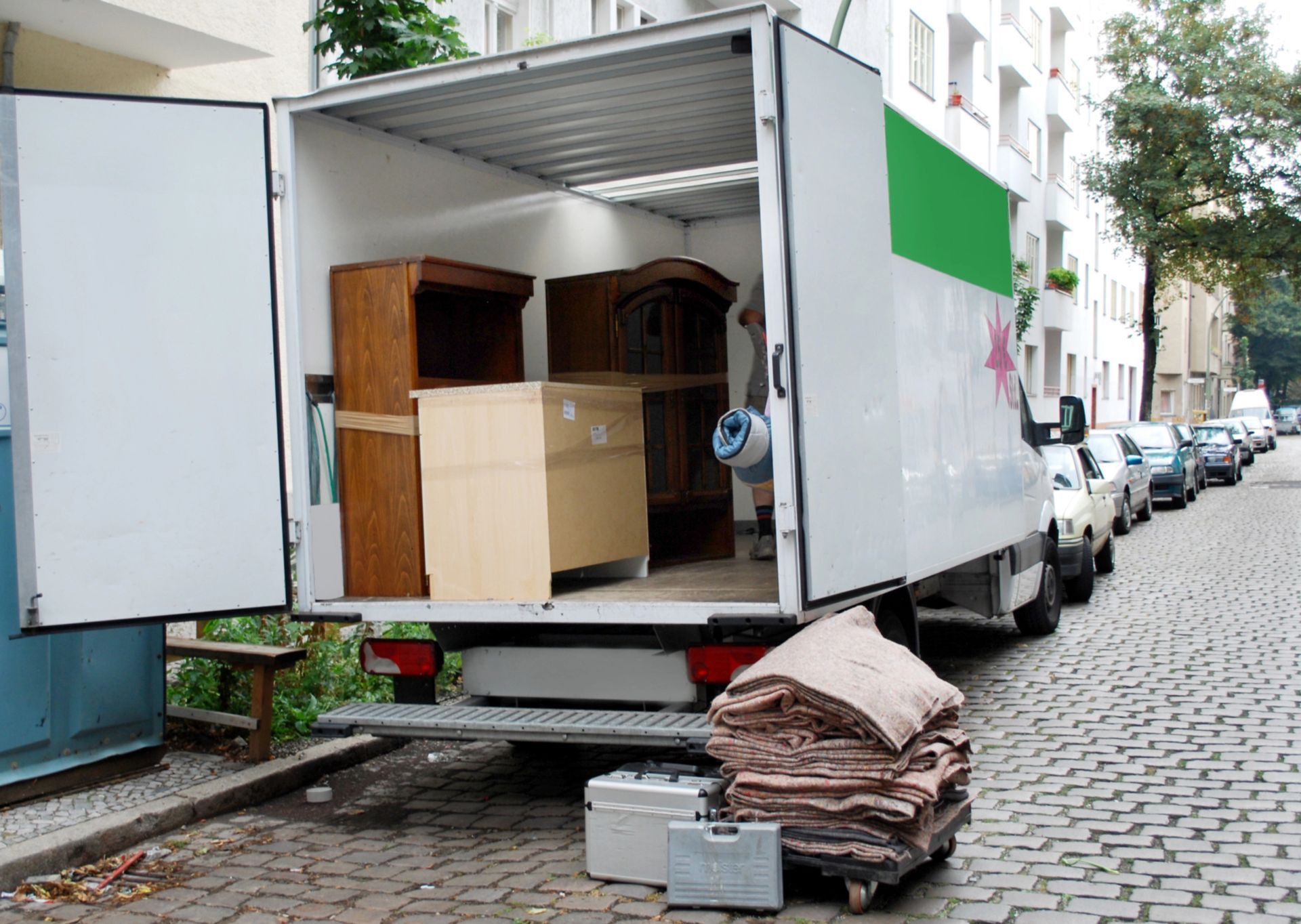 Camion per traslochi con cassone aperto parcheggiato su una strada acciottolata accanto a scatole e mobili impilati.
