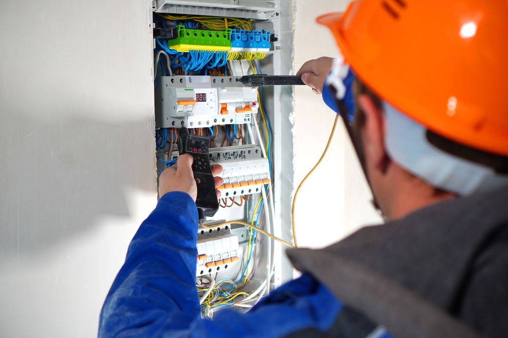 Elettricista che ispeziona un quadro elettrico aperto con i suoi attrezzi, indossando un elmetto arancione e una giacca blu.