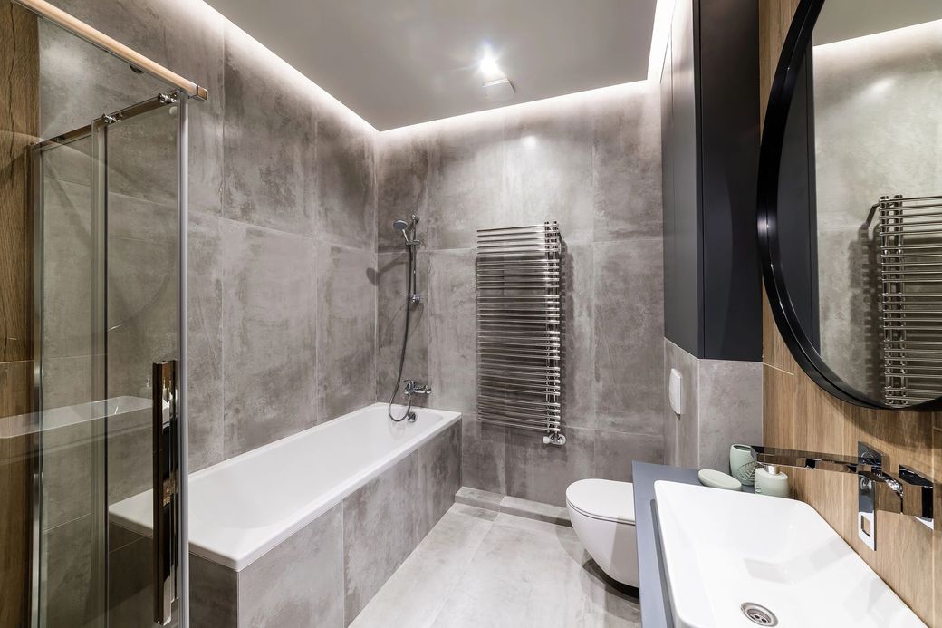 Bagno moderno con piastrelle grigie, vasca da bagno, doccia in vetro, scaldasalviette e specchio rotondo.