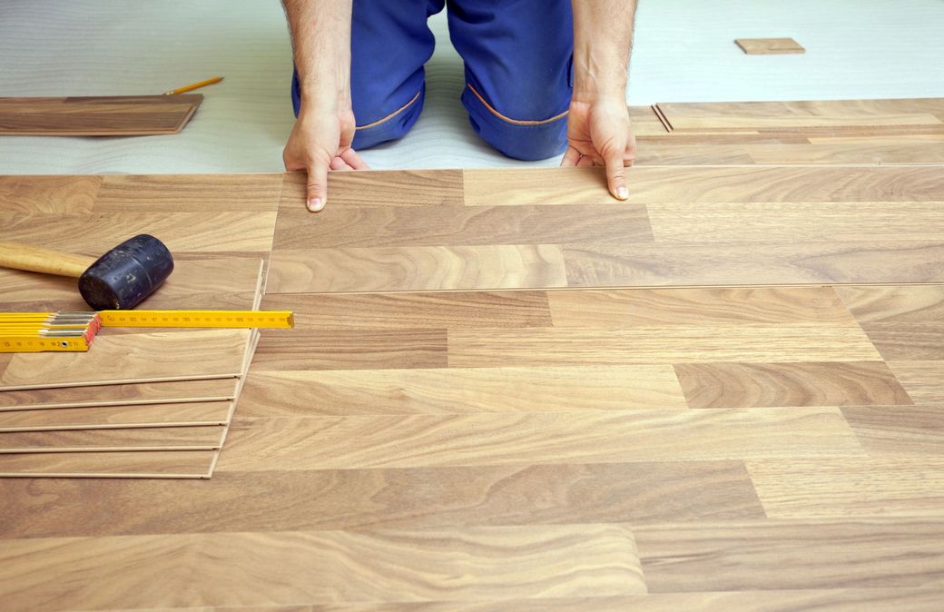 Una persona inginocchiata su un pavimento di legno misura e allinea le assi del pavimento con un righello e un martello.