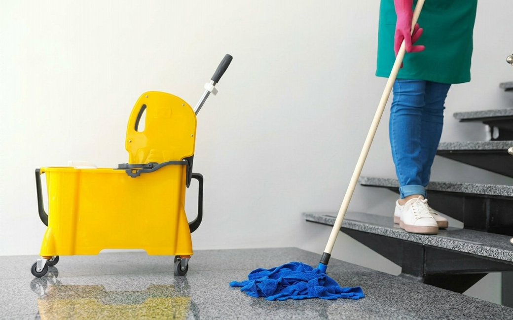 Una persona sta lavando un pavimento accanto a un secchio giallo e a dei prodotti per la pulizia vicino alle scale.