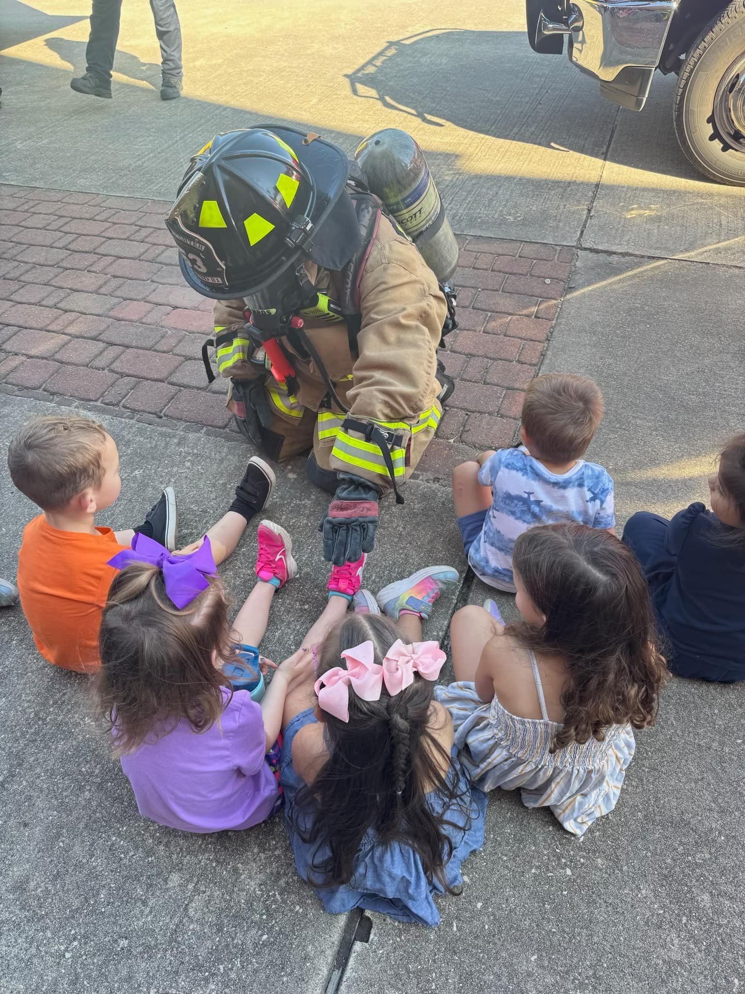 Firefighter kneeling with children outdoors, showing a black helmet and bright yellow gear.