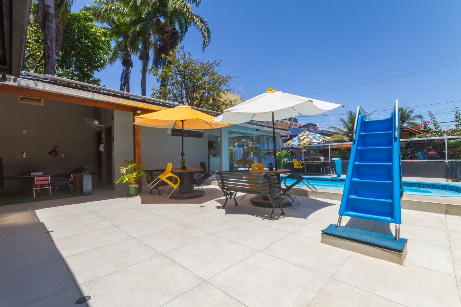 Um pátio com mesas, cadeiras, guarda-sóis e um escorregador ao lado de uma piscina.