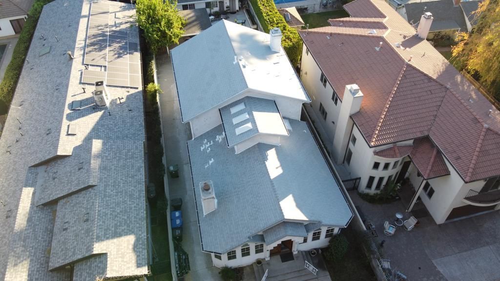 Aerial view of homes with light grey and brown roofs, greenery, and a narrow driveway.