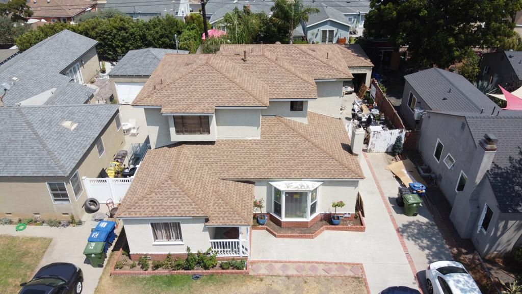 Aerial view of a light-colored house with a brown tile roof. Cars parked in front, neighboring homes visible.