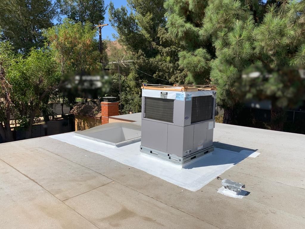 HVAC unit on a flat rooftop with a skylight, chimney, and surrounding trees under a bright blue sky.