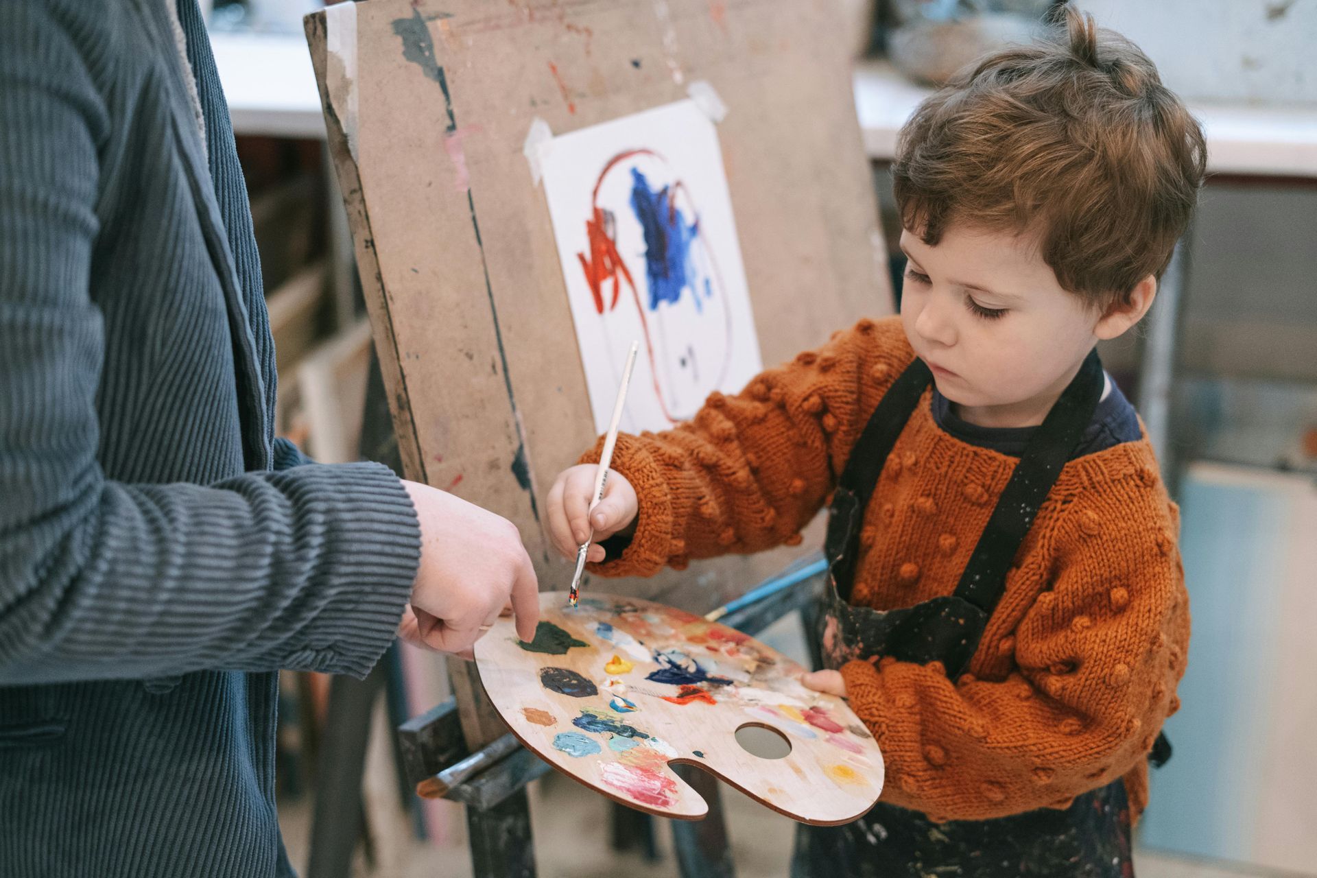 Child painting at easel with assistance; palette and paints are visible.