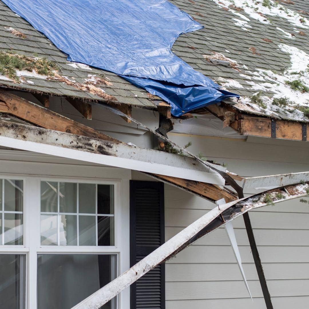 Severely damaged house roof with blue tarp and exposed broken beams above a window