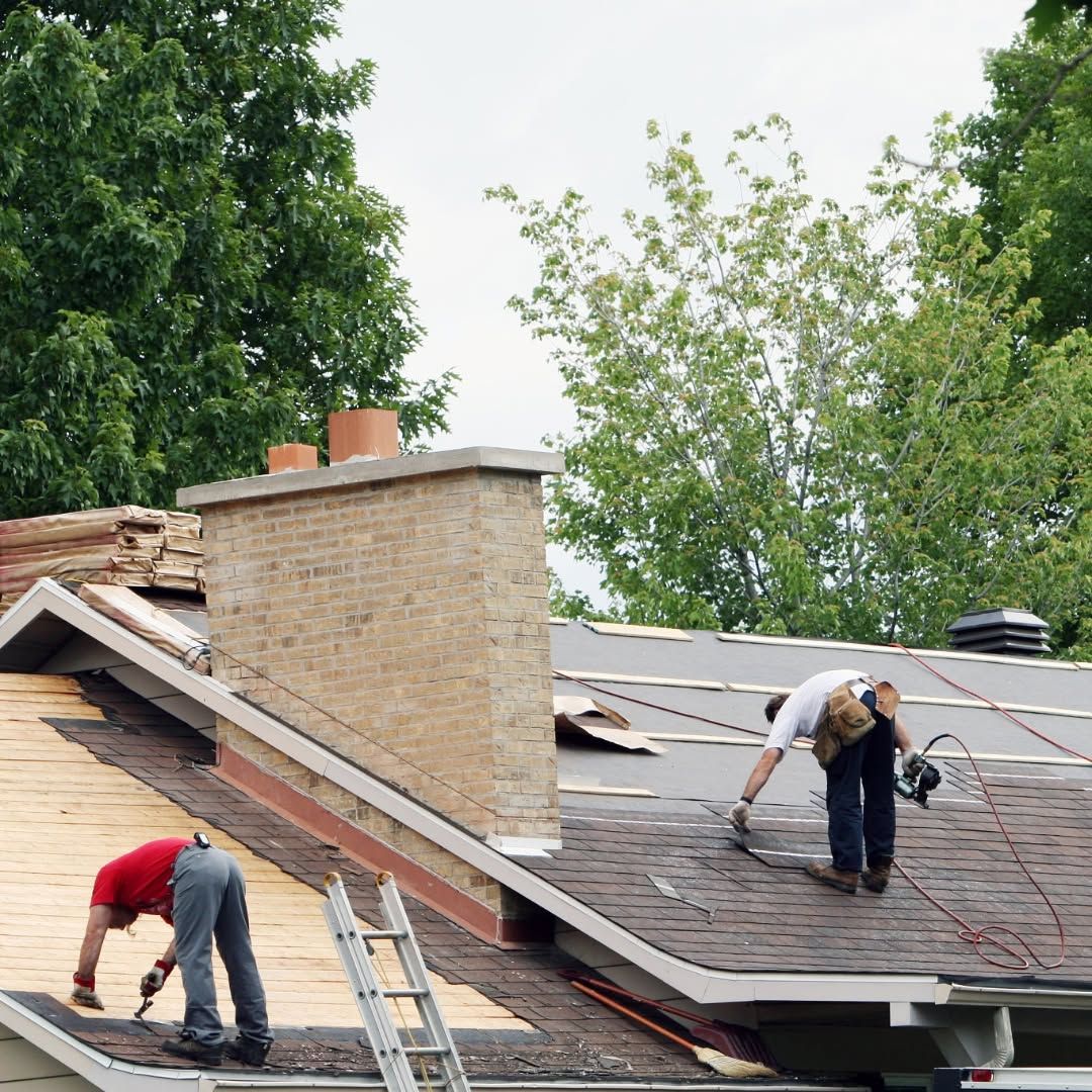 Two workers repairing a shingled roof beside a brick chimney on a house