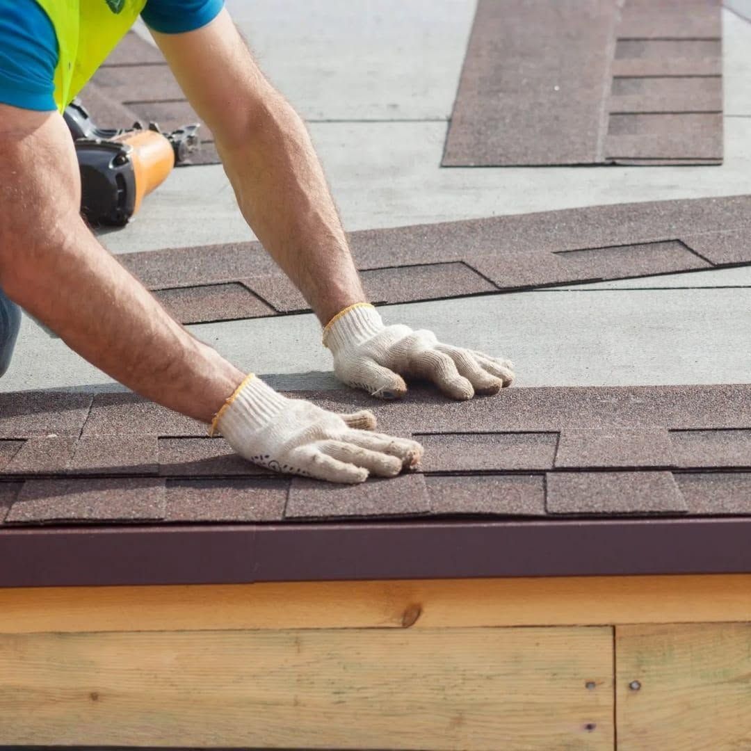 Worker installing brown roof shingles on a rooftop, hands pressing shingles into place.