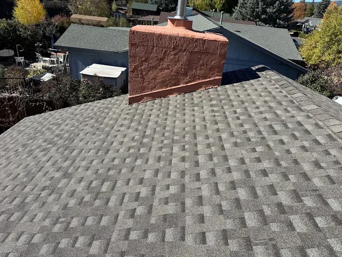 Rooftop with gray shingles and a large red brick chimney, overlooking trees and neighboring houses