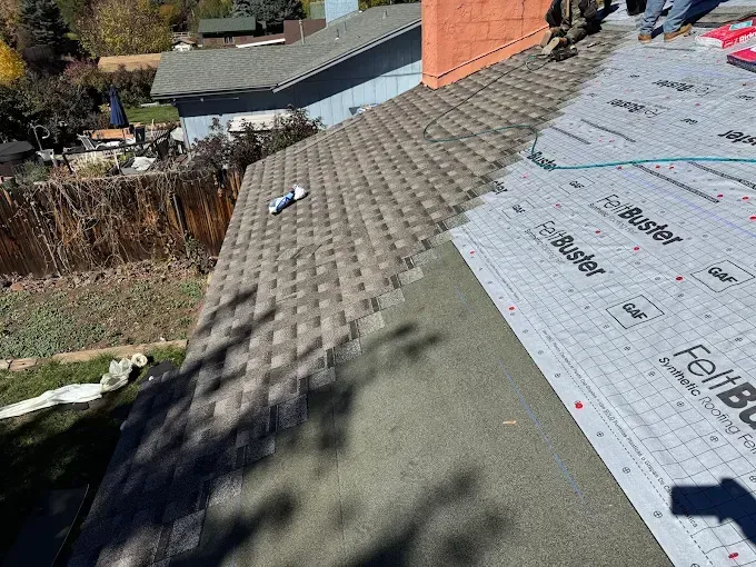 Rooftop with gray shingles, roofing underlayment, and a small tool near a chimney in sunlight