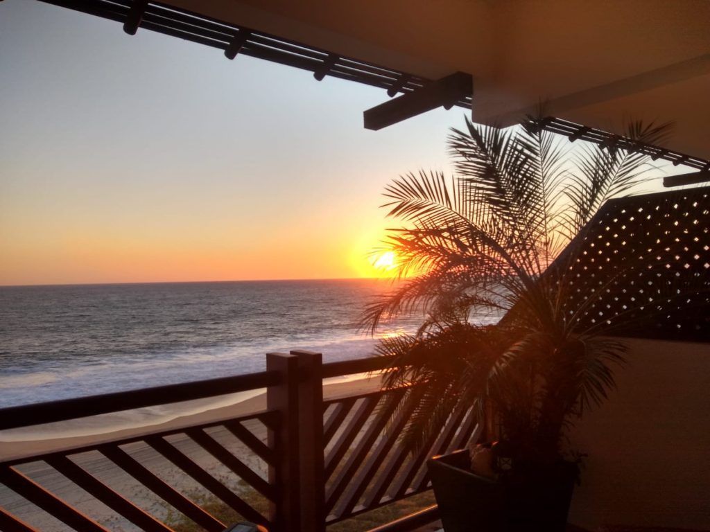 Sunset over ocean viewed from a balcony with palm tree and wooden railing.