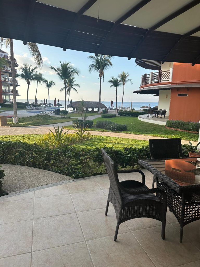 View from a patio with wicker furniture overlooking a beach with palm trees and a blue sky.