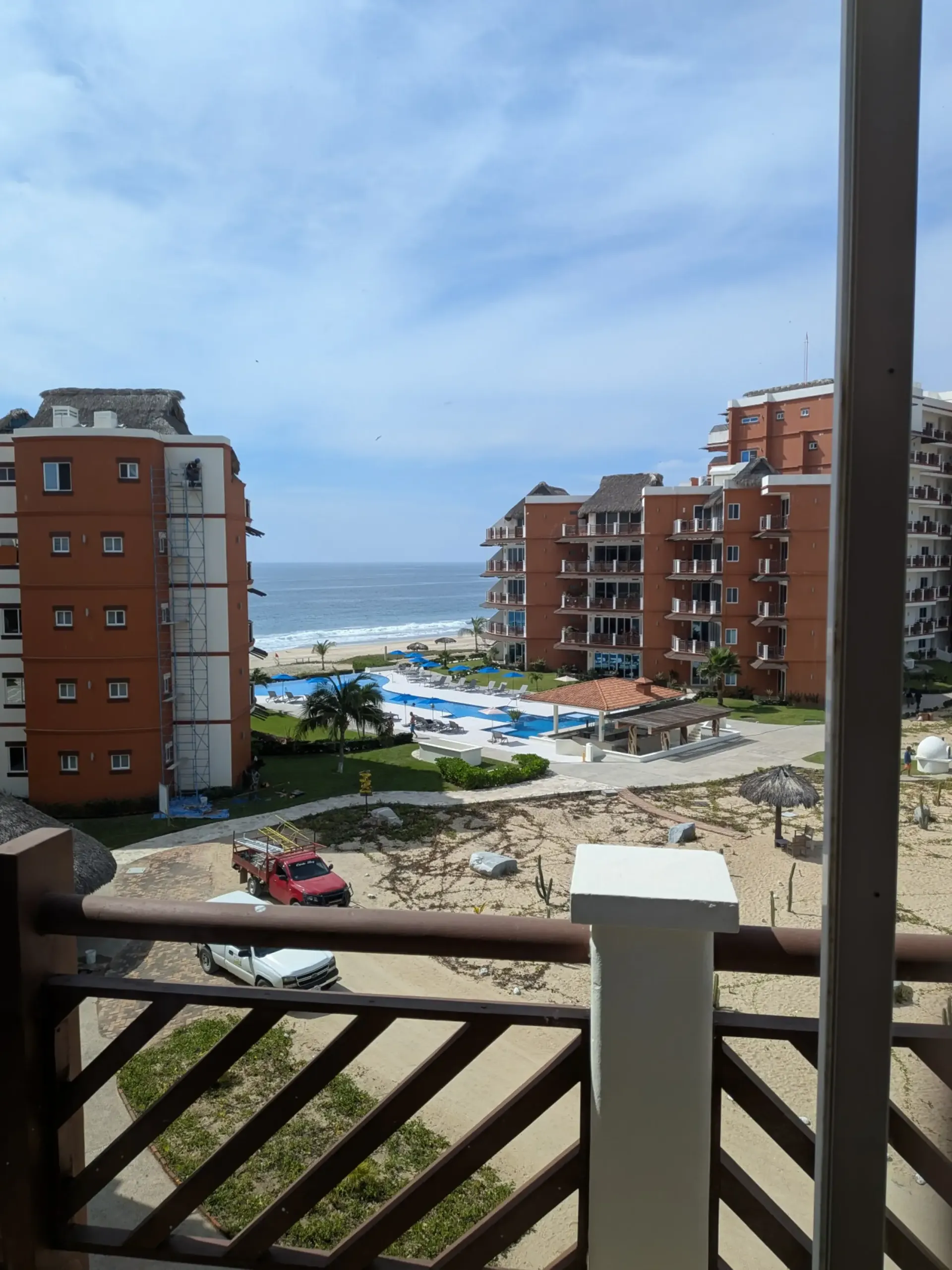 Ocean-view balcony overlooking apartment buildings, a pool, beach, and waves under a blue sky.